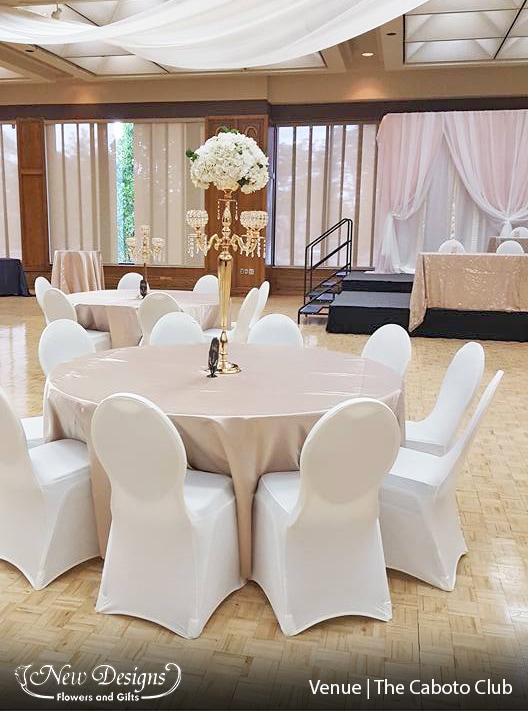 A crisp hall of white linens, white fitted spandex chaircovers, toned slightly with neutral blush satin tablecloths. A 5 posted gold and crystal candelabra is topped with a full centerpiece of Hydrangeas and Roses.